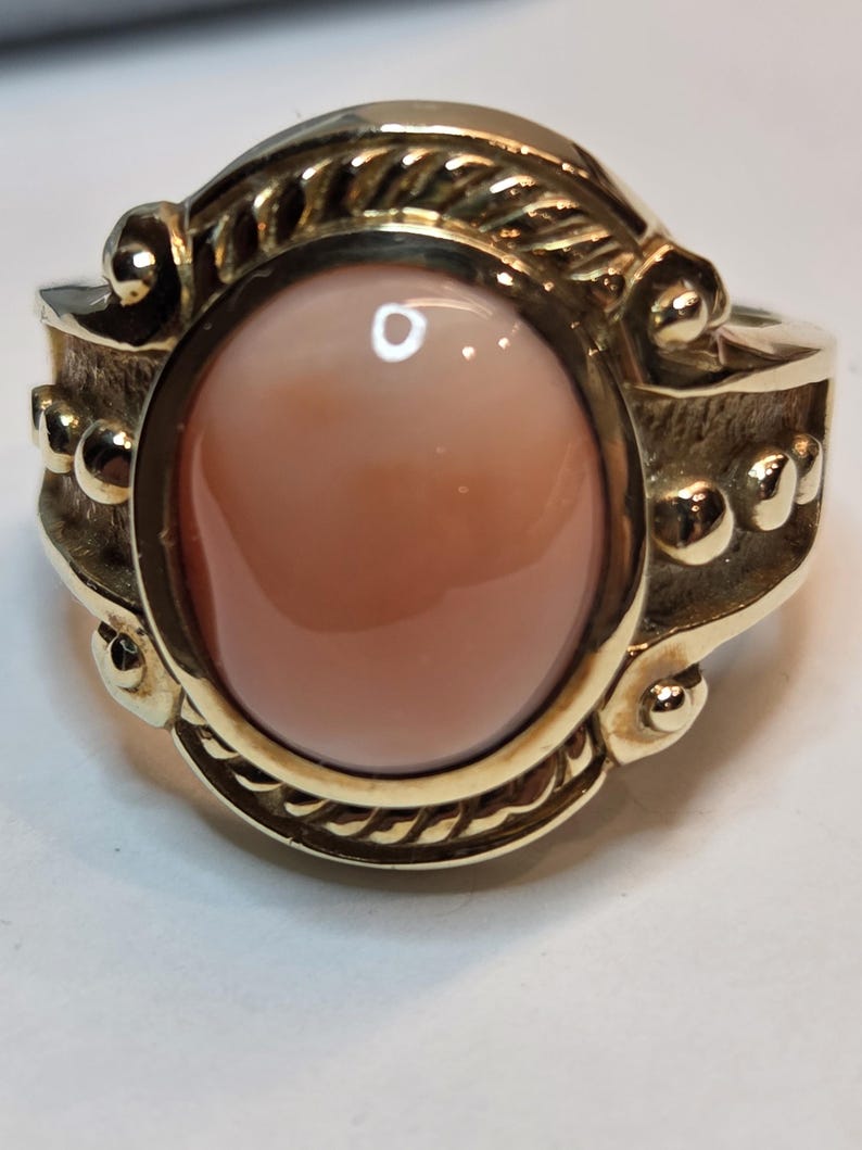 May include: A gold ring with an oval-shaped, light pink gemstone set in a rope-like design.