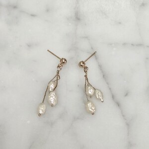 May include: A pair of gold-toned earrings with a post back. Each earring features a small gold ball at the top, with a delicate chain suspending three white, oblong pearls. The earrings are displayed on a white and gray marble surface.