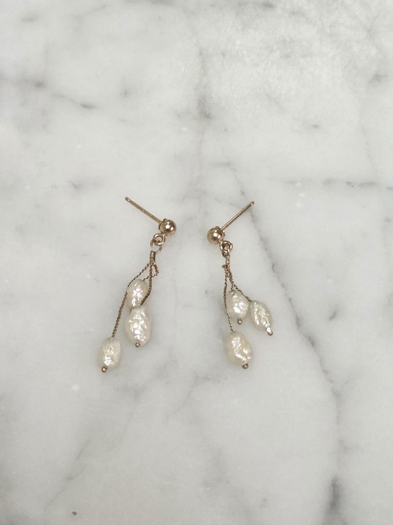 May include: A pair of gold-toned earrings with a post back. Each earring features a small gold ball at the top, with a delicate chain suspending three white, oblong pearls. The earrings are displayed on a white and gray marble surface.