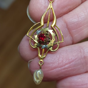 May include: A gold pendant with a red gemstone and a white pearl drop. The pendant is in an intricate, ornate design.