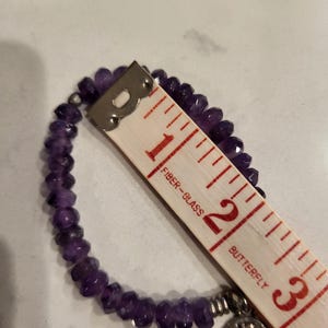 May include: A purple amethyst beaded bracelet with a silver charm featuring a butterfly design. The bracelet measures approximately 2 inches in diameter.
