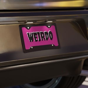 May include: A license plate with the word "WEIRDO" in white, stylized lettering against a bright pink background. The plate is framed in black and mounted on a vehicle.