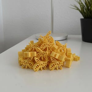 Op de afbeelding: Een stapel gele, bijvormige pasta op een wit oppervlak. De pasta, een lichtgele kleur, lijkt gemaakt te zijn van durumtarwe. De bijen zijn gedetailleerd met vleugels en lichaamssegmenten. Een kleine potplant staat op de achtergrond.