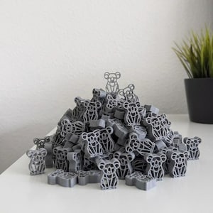 Op de afbeelding: Een grote stapel grijze koala-vormige objecten. Elke koala heeft een gedetailleerd uitgesneden ontwerp. De koala's zijn verspreid op een wit oppervlak, met een kleine plant in een pot op de achtergrond.