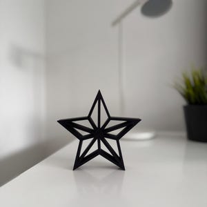 Könnte beinhalten: Ein schwarzes, fünfzackiges sternförmiges Dekorationsobjekt. Der Stern hat ein geometrisches Design mit inneren Linien, das einen dreidimensionalen Effekt erzeugt. Es steht auf einer weißen Oberfläche, mit einer Lampe und einer Pflanze im Hintergrund.