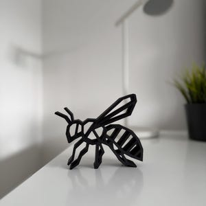 May include: A black geometric bee sculpture with a modern design. The bee is made of solid black material and has a detailed, angular structure. The sculpture is placed on a white surface.