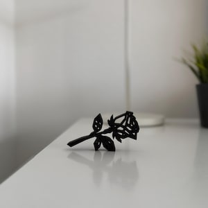 Peut inclure: Une sculpture de rose noire repose sur une surface blanche. La rose est finement conçue avec des détails découpés, montrant les pétales et les feuilles. La sculpture est inclinée, avec la tige pointant vers la gauche. Le fond est blanc flou.