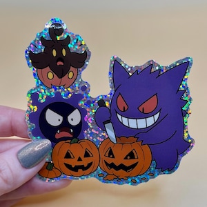 May include: A holographic sticker featuring cartoon characters, including a purple Gengar holding a knife, a black Gastly, and a brown and orange character. Two carved orange pumpkins are also featured. The sticker has a glittery, iridescent border.
