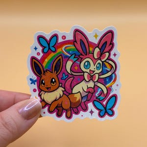 May include: A colorful sticker featuring two cartoon characters, Eevee and Sylveon, with a rainbow and butterflies. Eevee is brown and Sylveon is pink and white. The sticker has a white border and is held by a hand.