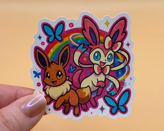 May include: A colorful sticker featuring two cartoon characters, Eevee and Sylveon, with a rainbow and butterflies. Eevee is brown and Sylveon is pink and white. The sticker has a white border and is held by a hand.