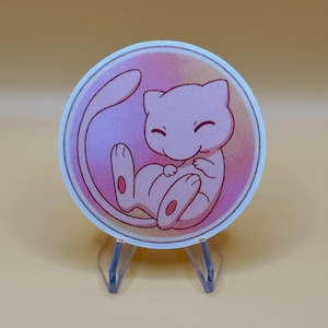May include: A round sticker featuring a pink cartoon character with a long tail, curled up in a fetal position. The character has a smiling face and is set against a gradient background of pink and orange. The sticker is displayed on a clear plastic stand.