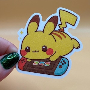 May include: A yellow Pikachu sticker with a glittery texture. The sticker features Pikachu playing a video game on a brown and blue game console. The sticker has a white outline and a light orange background.