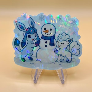 May include: A holographic sticker featuring a winter scene with cartoon characters. The sticker shows a snowman with a blue scarf, and two cartoon creatures in shades of blue. Snowflakes are scattered around the characters.