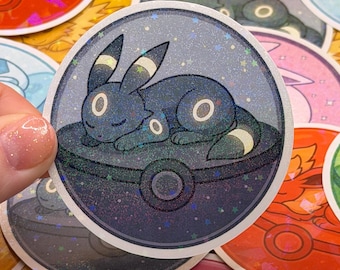 May include: A collection of colorful, circular stickers featuring various cartoon creatures. The primary sticker shows a dark blue Umbreon character resting inside a dark blue and gray Poke Ball, with a glittery background.