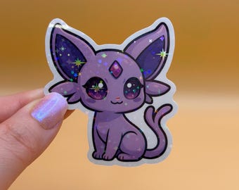 May include: A holographic sticker featuring a purple cartoon cat with large eyes and a gem on its forehead. The cat has large ears and a curled tail. The sticker has a white border and is held against a light orange background.