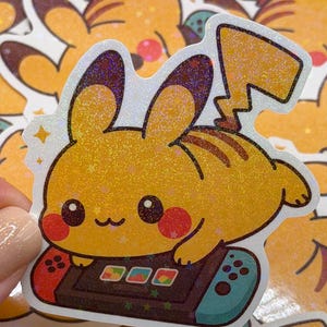 May include: A yellow Pikachu sticker with a holographic finish. The sticker features Pikachu playing a video game on a Nintendo Switch-style controller. The sticker has a white border and is surrounded by other similar stickers.