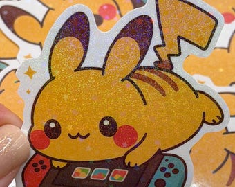 May include: A yellow Pikachu sticker with a holographic finish. The sticker features Pikachu playing a video game on a Nintendo Switch-style controller. The sticker has a white border and is surrounded by other similar stickers.