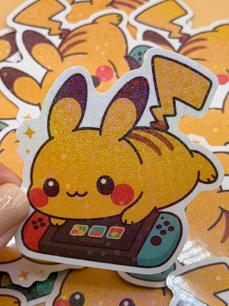 May include: A yellow Pikachu sticker with a holographic finish. The sticker features Pikachu playing a video game on a Nintendo Switch-style controller. The sticker has a white border and is surrounded by other similar stickers.