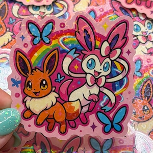 May include: A holographic sticker featuring a brown Eevee and a pink Sylveon character standing in front of a colorful rainbow. The design includes blue butterflies, small pink circles, and sparkling star accents on a light pink background.