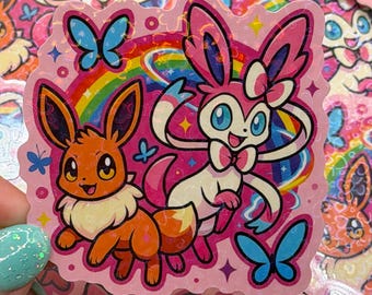 May include: A holographic sticker featuring a brown Eevee and a pink Sylveon character standing in front of a colorful rainbow. The design includes blue butterflies, small pink circles, and sparkling star accents on a light pink background.