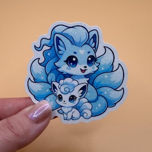 May include: A sticker featuring a cartoon illustration of two foxes. The larger fox is light blue with multiple fluffy tails and a heart-shaped marking. The smaller fox is white. Both have large, expressive eyes and are outlined in white.