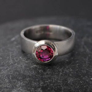May include: A silver ring featuring a wide band and a bezel-set, round, deep pink gemstone. The ring is presented on a dark, textured surface, accentuating the contrast between the metal and the stone.