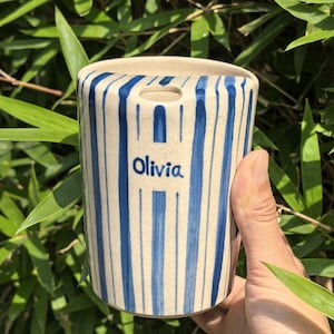 May include: A cylindrical ceramic vase with a cream-colored base and vertical blue stripes. The word "Olivia" is written in blue on the front. The vase has a small opening at the top.