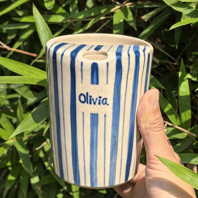 May include: A cylindrical ceramic vase with a cream-colored base and vertical blue stripes. The word "Olivia" is written in blue on the front. The vase has a small opening at the top.