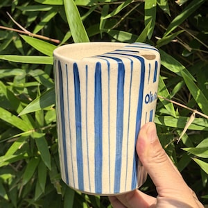 May include: A cylindrical ceramic vase with vertical blue stripes on a cream background. The word "Olivia" is written in blue on the side. The vase is held by a hand against a backdrop of green bamboo leaves.