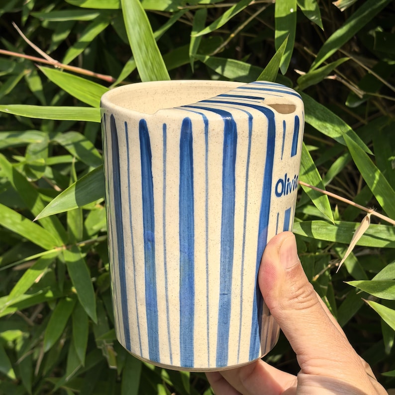 May include: A cylindrical ceramic vase with vertical blue stripes on a cream background. The word "Olivia" is written in blue on the side. The vase is held by a hand against a backdrop of green bamboo leaves.