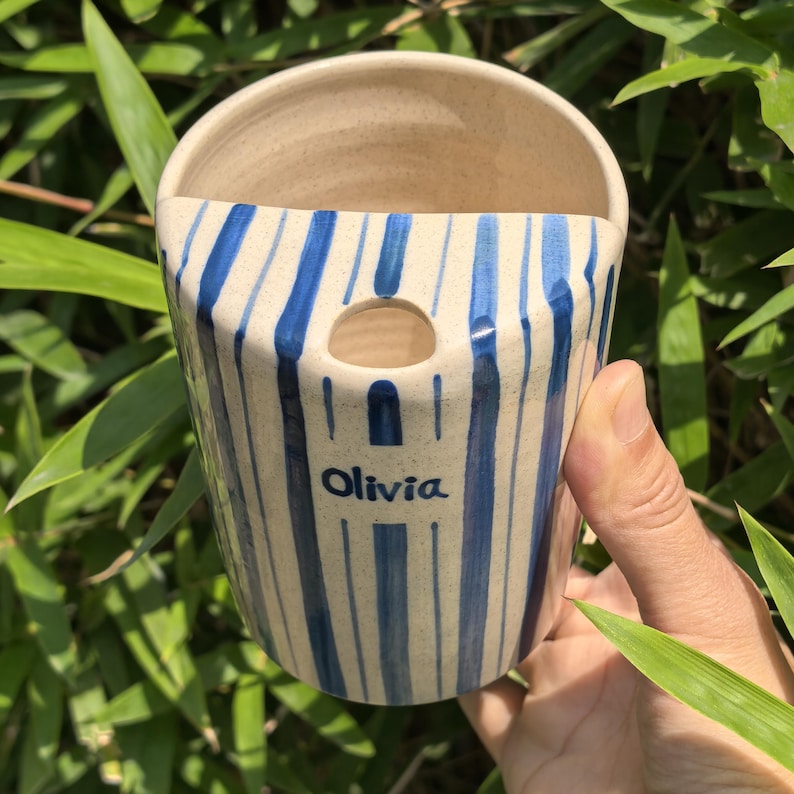 May include: A ceramic wall pocket with a cream-colored interior and exterior. The exterior features vertical blue stripes. The name "Olivia" is written in blue on the front. The pocket is held by a hand against a background of green foliage.