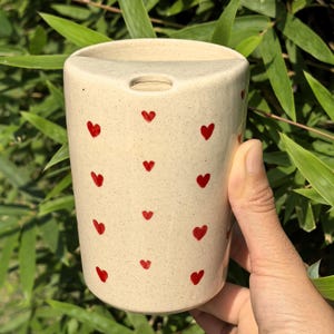 Può includere: Un vaso in ceramica beige con una parte superiore triangolare e un piccolo foro. Il vaso è decorato con piccoli cuori rossi dipinti a mano. Lo sfondo è un'immagine sfocata di foglie verdi.