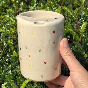 May include: A beige ceramic toothbrush holder with a cylindrical shape and a curved top. The holder is decorated with small, colourful heart-shaped designs. The holder is being held in a hand.