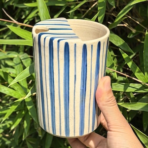 May include: A cylindrical ceramic mug with a lid, featuring vertical blue stripes against a cream-colored background. The mug has a unique cutout on the lid. The design is simple and elegant, suitable for beverages.