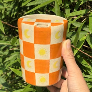 Op de afbeelding: Een cilindrische keramische vaas met een geblokt patroon van oranje en crèmekleurige vierkanten. De vaas is versierd met gele zon- en maanmotieven. De bovenkant van de vaas heeft een kleine rechthoekige opening.