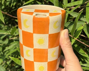 Puede incluir: Un jarrón cilíndrico de cerámica con un patrón a cuadros de cuadrados naranjas y crema. El jarrón está decorado con motivos de sol y luna amarillos. La parte superior del jarrón tiene una pequeña abertura rectangular.