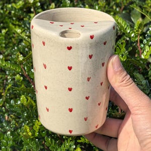 May include: A beige ceramic toothbrush holder with a speckled finish and small red heart designs scattered across the surface. The holder has a curved top and a hole for a toothbrush.