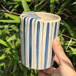 May include: A ceramic mug with a cream-colored base and vertical blue stripes. The mug has a unique shape with a cutout on the rim. The word "ia" is written on the side in cursive. The mug is held against a backdrop of green foliage.