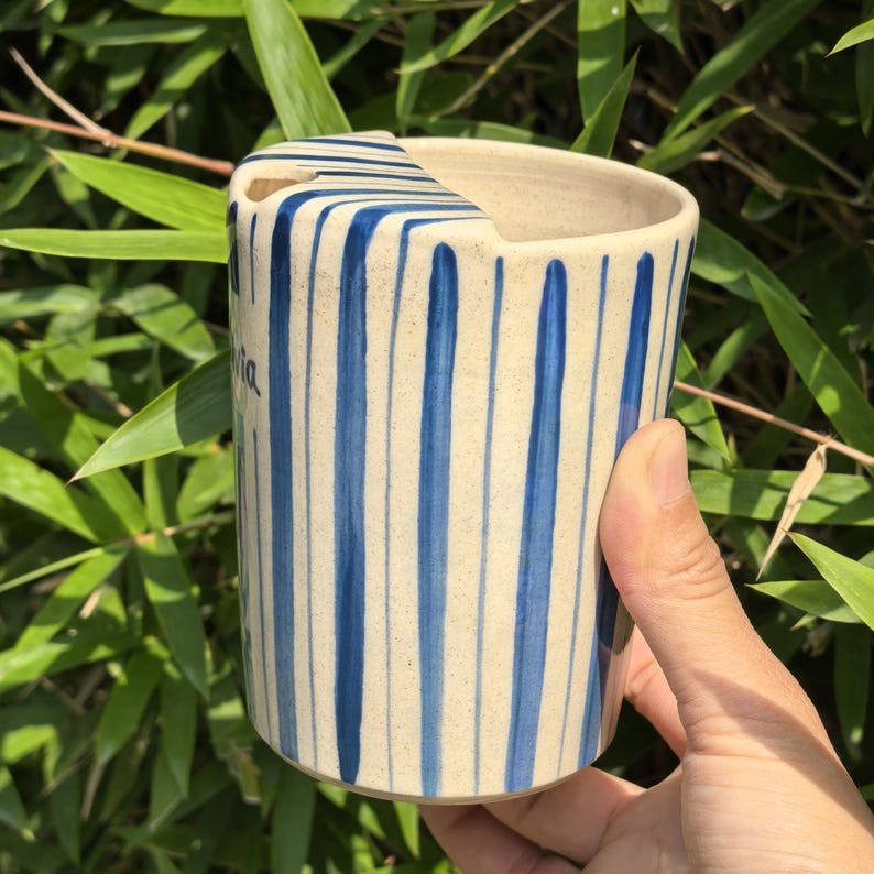 May include: A ceramic mug with a cream-colored base and vertical blue stripes. The mug has a unique shape with a cutout on the rim. The word "ia" is written on the side in cursive. The mug is held against a backdrop of green foliage.