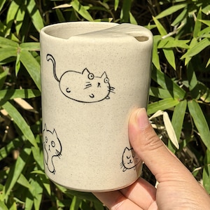 May include: A beige ceramic mug with a rounded top and a small opening. The mug is decorated with black line drawings of cartoon cats. The mug is held by a hand, with a green leafy background.