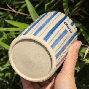 May include: A ceramic mug with a beige base and blue vertical stripes. The word "Olivia" is printed in blue on the side. The mug is held in a hand against a background of green foliage.