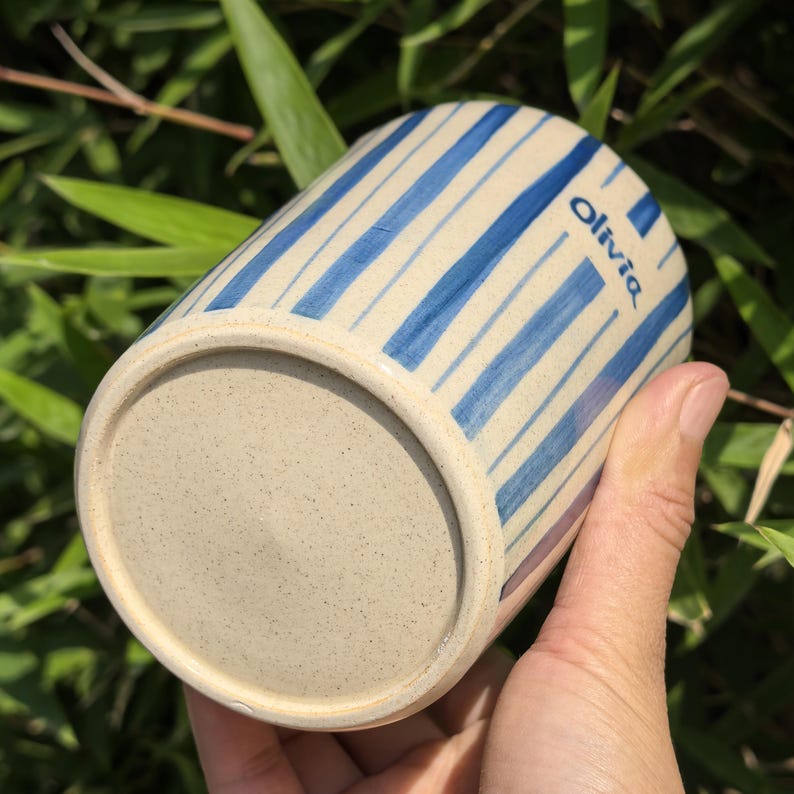 May include: A ceramic mug with a beige base and blue vertical stripes. The word "Olivia" is printed in blue on the side. The mug is held in a hand against a background of green foliage.