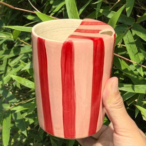 May include: A cylindrical ceramic mug with a pink base and vertical red stripes. The mug has a unique cut-out design on the rim. The mug is held in a hand against a backdrop of green foliage.