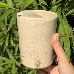 Peut inclure: Un gobelet en céramique beige avec un couvercle, présentant une texture subtilement mouchetée et un motif d'herbe peint. Le gobelet est présenté sur un fond de feuilles de bambou vertes.