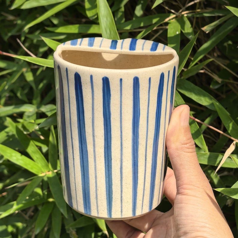 May include: A ceramic toothbrush holder with a cream-colored base and vertical blue stripes. The holder has a rectangular opening at the top and is held by a hand against a backdrop of green foliage.