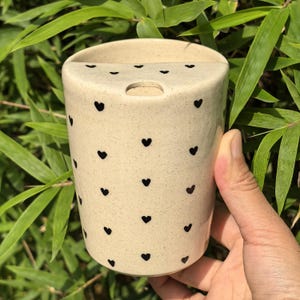 Könnte beinhalten: Beigefarbener Keramik-Pflanztopf mit schwarzen herzförmigen Mustern. Der Pflanztopf hat eine gewölbte Oberseite mit einem Loch. Das Design befindet sich vor einem Hintergrund aus grünen Blättern. Die Herzen sind gleichmäßig verteilt.