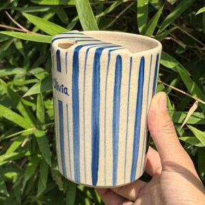 May include: A ceramic toothbrush holder with a cream-colored base and vertical blue stripes. The name "Olivia" is written in blue script on the side. The holder has a curved top with a slot for toothbrushes.