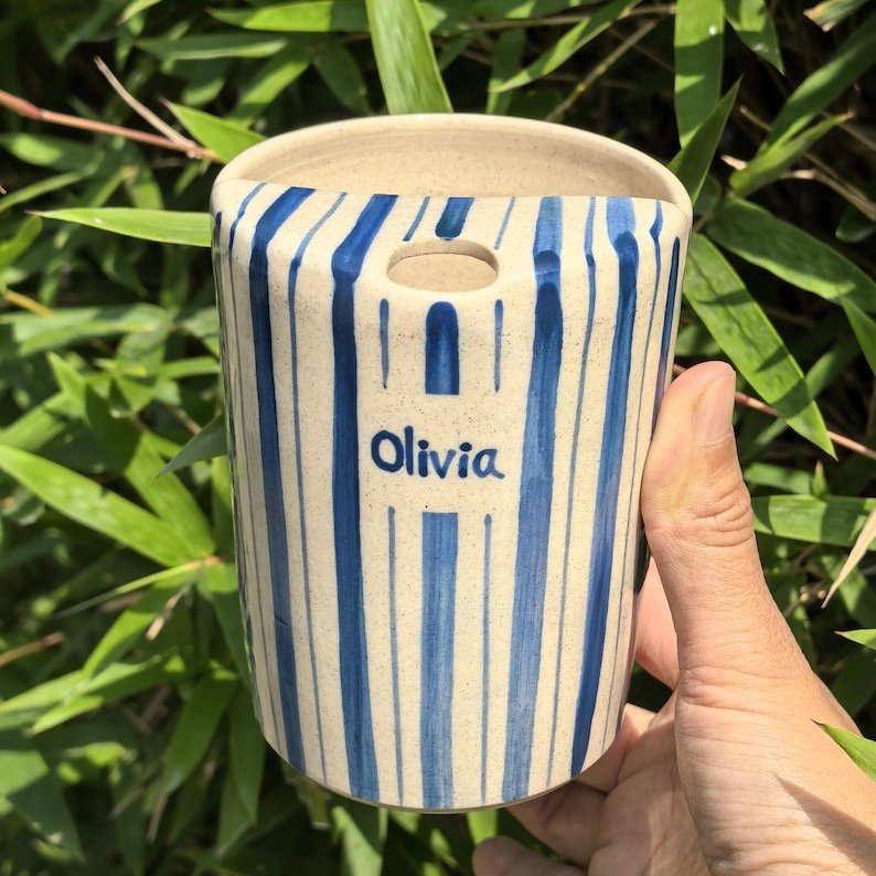 May include: A ceramic wall pocket with a cream-colored base and blue vertical stripes. The name "Olivia" is written in blue script on the front. The pocket has a hole at the top for hanging and is held by a hand against a backdrop of green foliage.