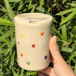 Könnte beinhalten: Ein beigefarbener Zahnbürstenhalter aus Keramik mit zylindrischer Form. Der Halter ist mit bunten Herzformen in Gelb, Orange, Rot, Grün und Blau verziert. Er hat eine gewölbte Oberseite mit einem Loch für die Zahnbürste.