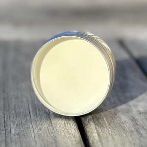 May include: An open silver metal tin filled with a pale yellow, creamy product. The tin is positioned on a weathered wooden surface. The contents suggest a skincare or cosmetic product, possibly a balm or cream.